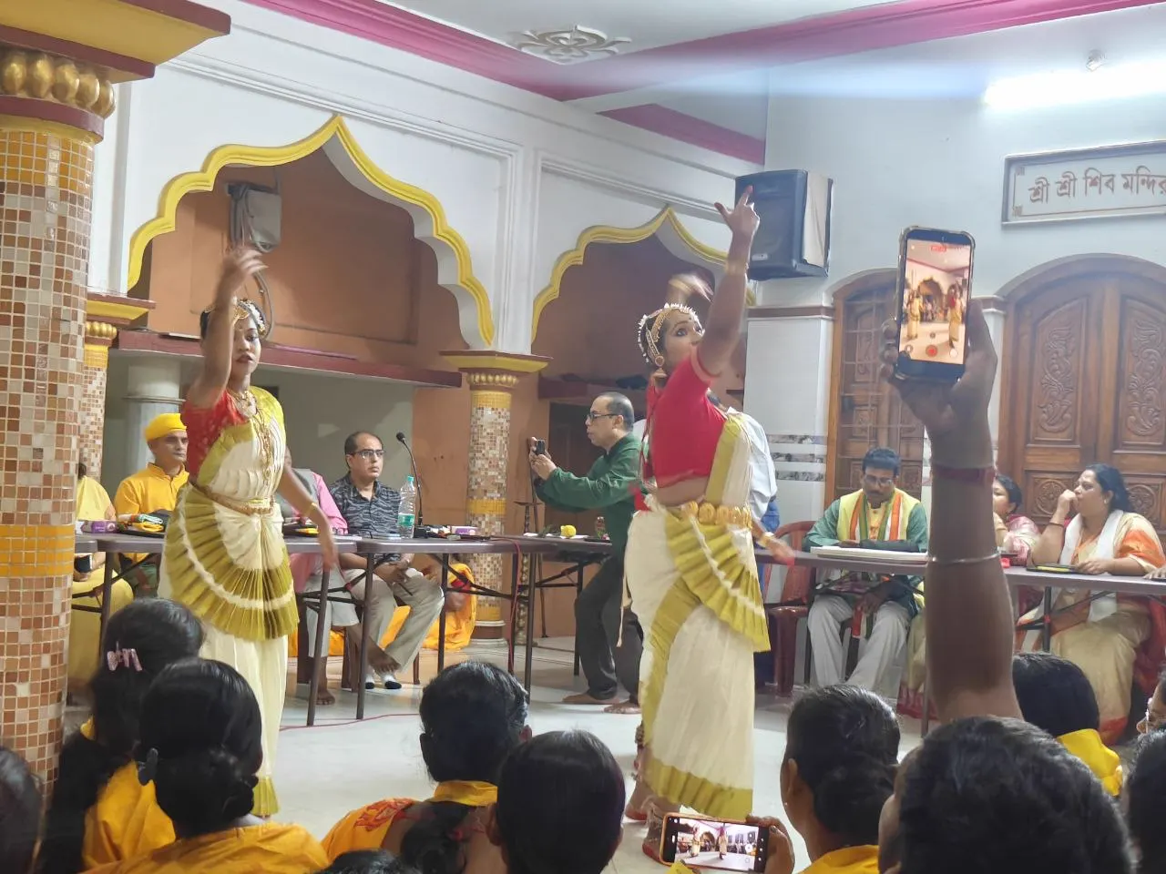 Two dancers in cream, red and gold perform in the hall of Sri Sri Shiv Mandir while the gathering watches.