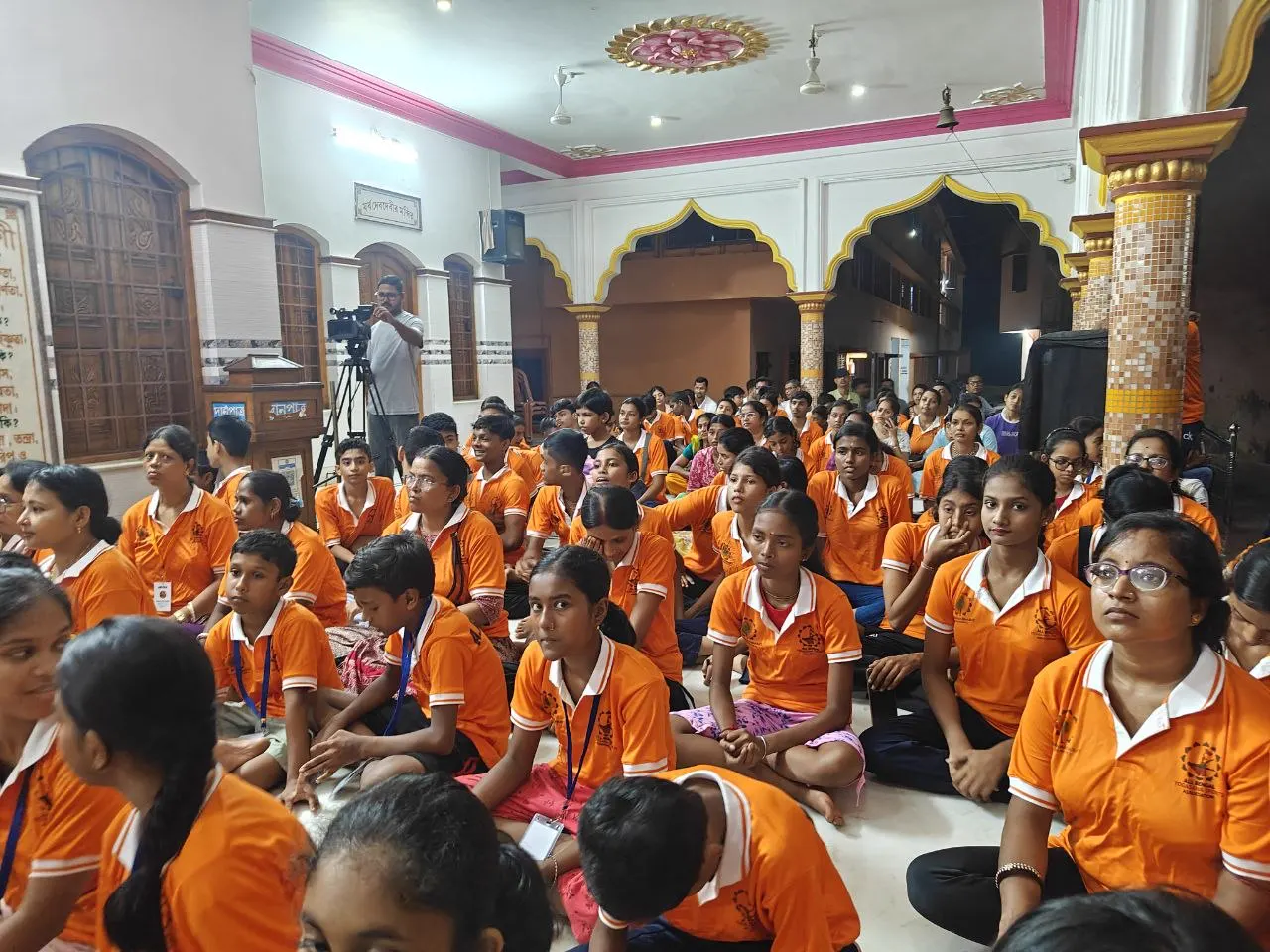 Athletes in orange shirts sit on the floor of the hall, facing the front.