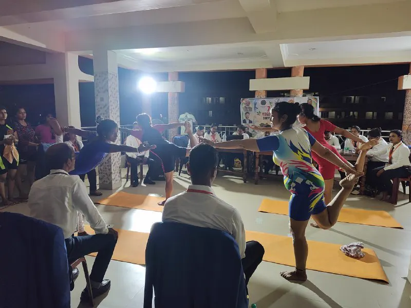 An athlete holds a standing balance on a mat at night, with judges seated behind.