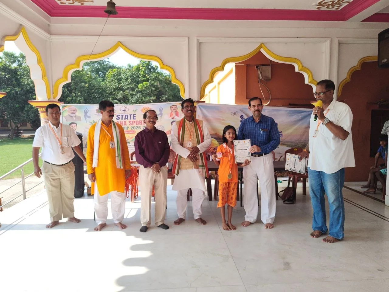 Officials stand with an athlete in orange as a certificate is held in the hall.