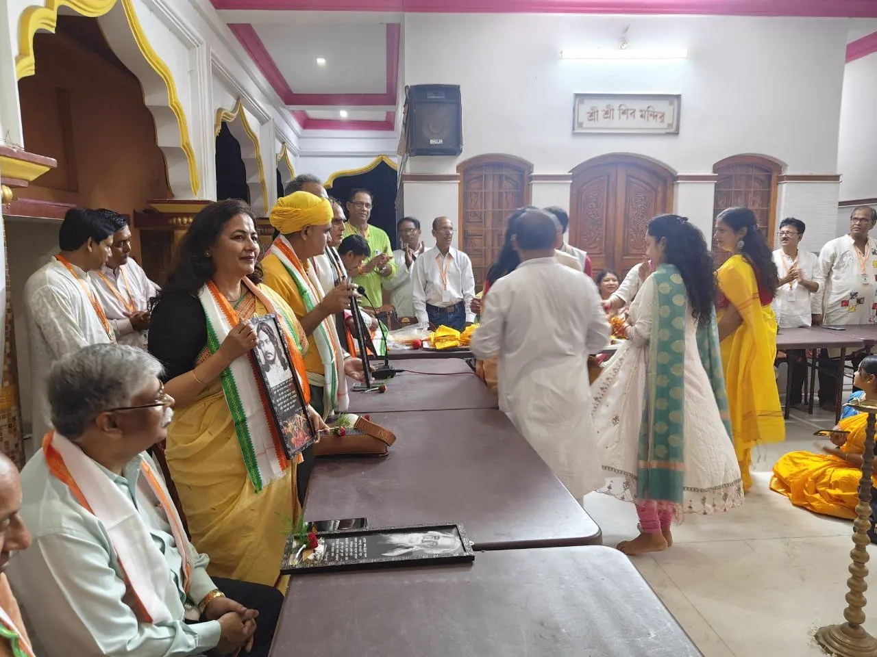 A woman in a yellow saree holds a framed portrait of Rabindranath Tagore at the long table, with a second portrait lying on the table.